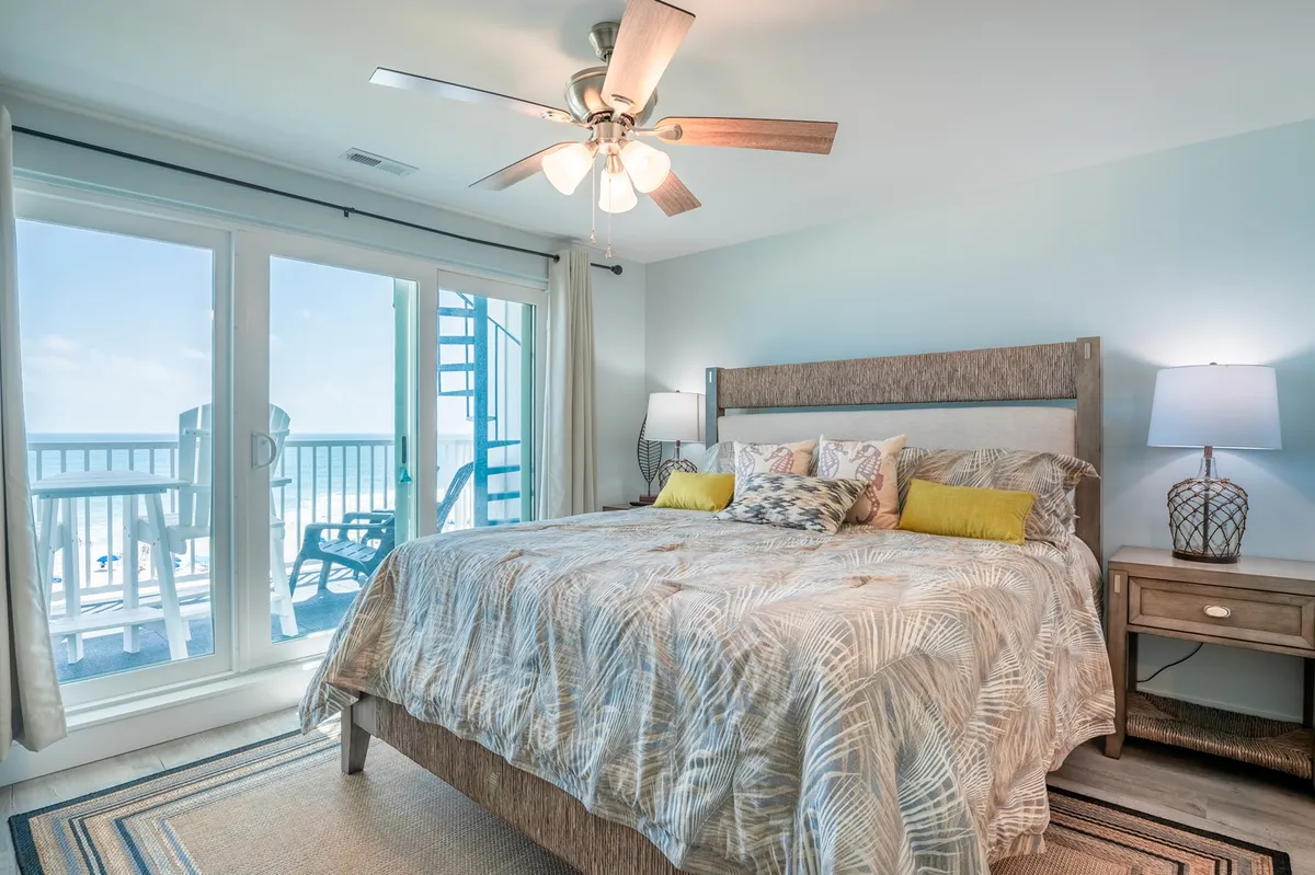 A serene master bedroom with a king-sized bed topped with decorative pillows. Large windows let in ample natural light and offer a sweeping ocean view. A ceiling fan and bedside tables add comfort and convenience. The layout includes easy access to a private balcony, visible through the glass doors.