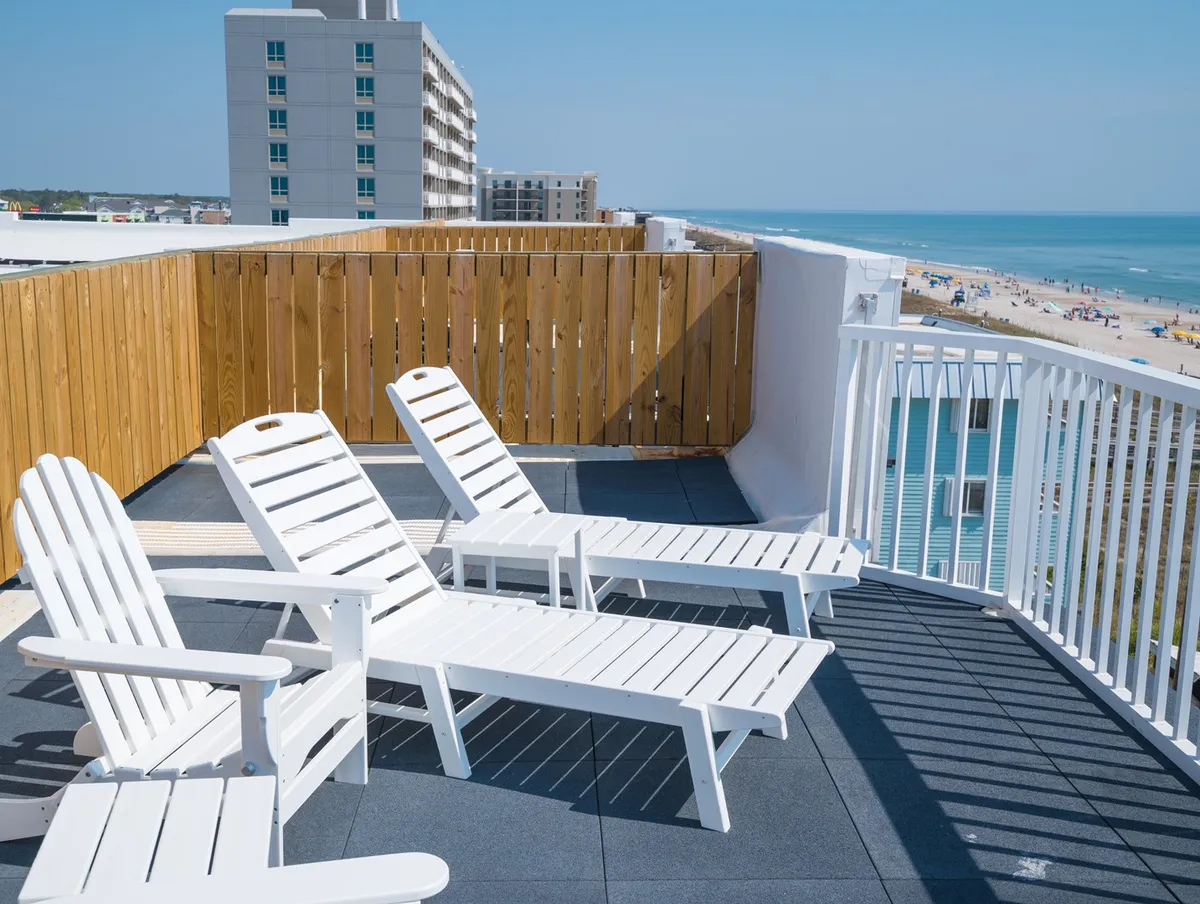 The exterior hero image showcases a serene oceanfront view framed by a wooden fence, with inviting white lounge chairs positioned to soak in the sunrise or unwind under the stars. The tranquil atmosphere suggests relaxation and leisure, all set against the stunning Carolina Beach shoreline.