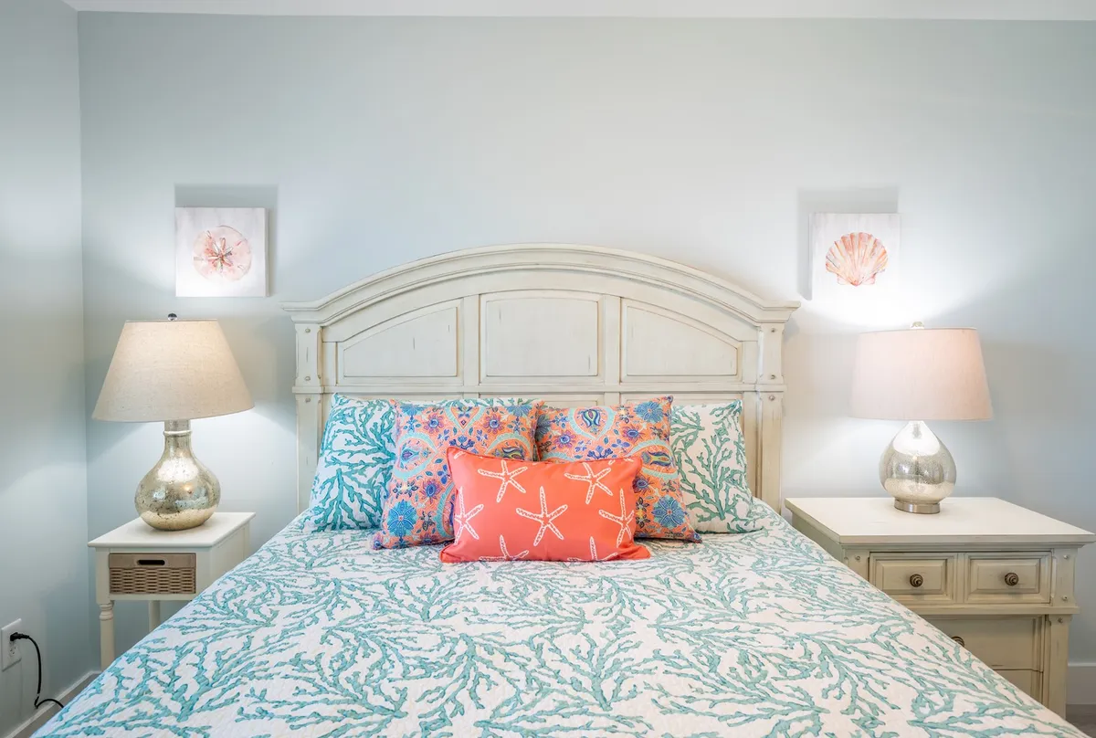 A guest bedroom with a comfortable bed adorned with decorative pillows, nightstands with lamps, and shell art on the walls, offering a cozy and soothing environment. The overall mood is restful and inviting, ideal for unwinding after a busy day.