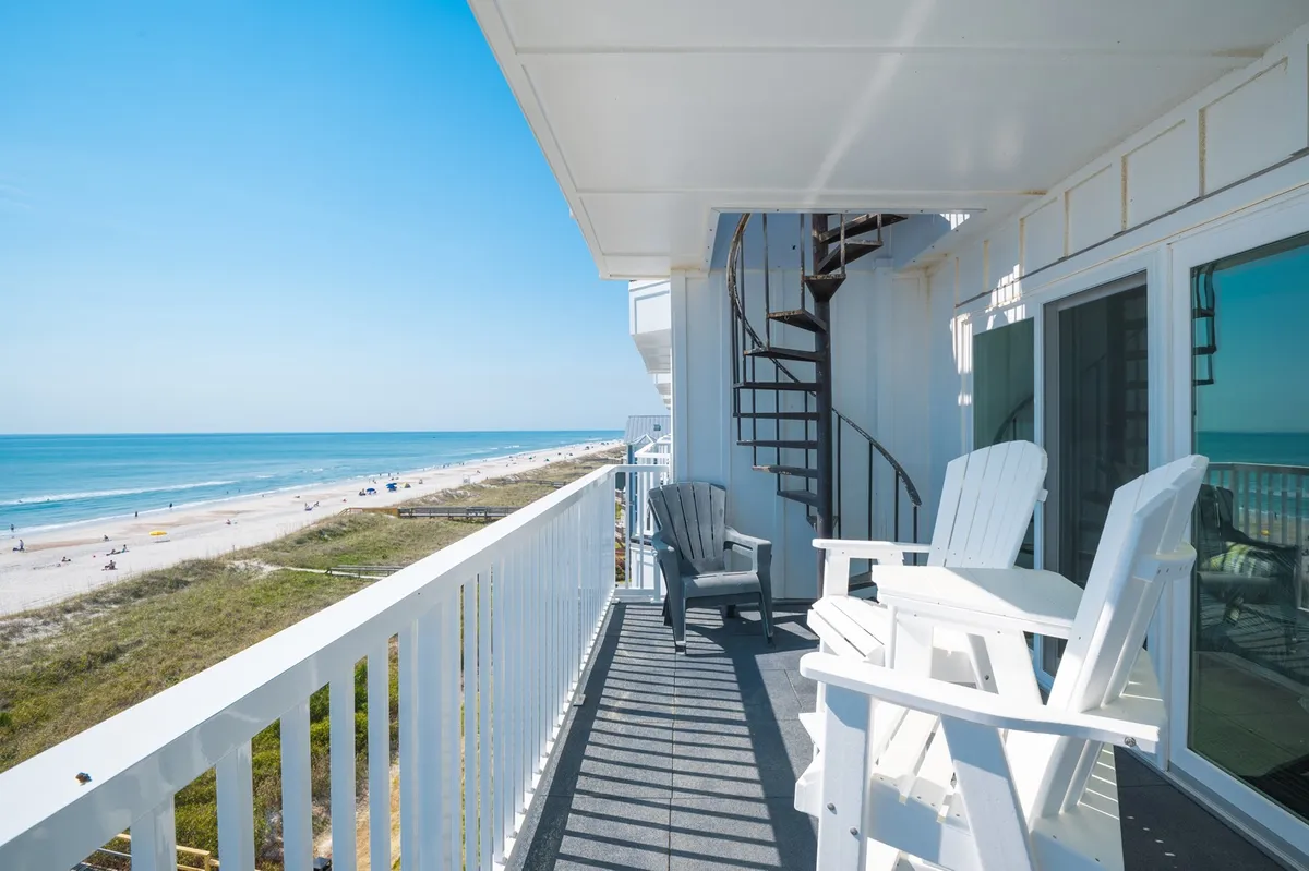 The photo shows a peaceful rooftop patio featuring white Adirondack chairs, a spiral staircase leading upward, and sweeping views of the beach and ocean beyond. The elevated vantage point provides a unique perspective for enjoying sunsets, stargazing, or private gatherings.