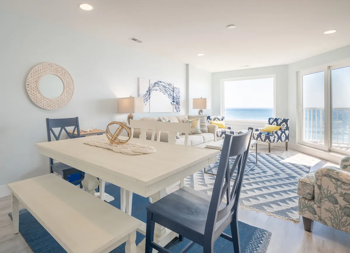 An open dining area with a large table set for six, surrounded by comfortable chairs. Expansive windows reveal breathtaking sea views, and a cozy seating area nearby enhances the sense of communal space.