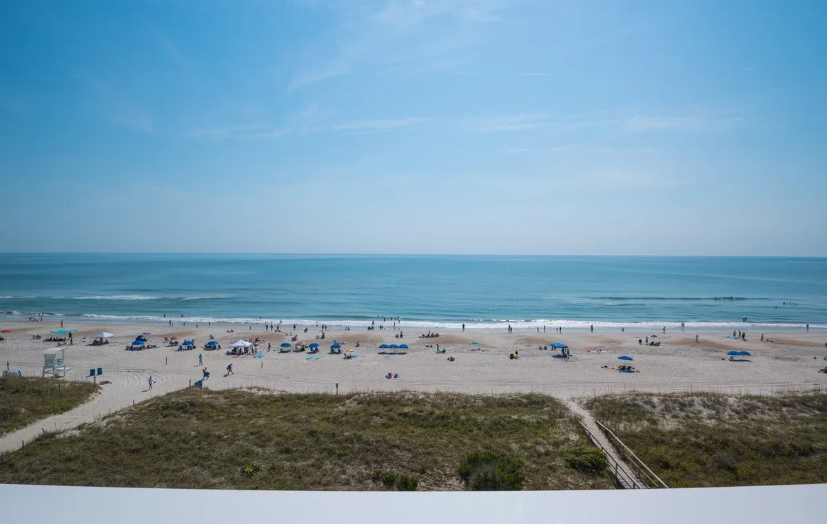 A vibrant view of the beach, with people sunbathing, swimming, playing beach games, and relaxing under colorful umbrellas. The ocean stretches to the horizon, evoking the energy and joy of time spent in nature.