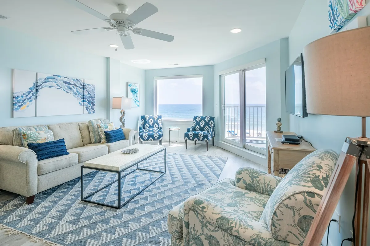 A bright and airy living room featuring a plush sofa, coffee table, decorative accent chairs, and a large window framing a sweeping ocean view. A lamp and modern decor add to the inviting, contemporary ambiance.