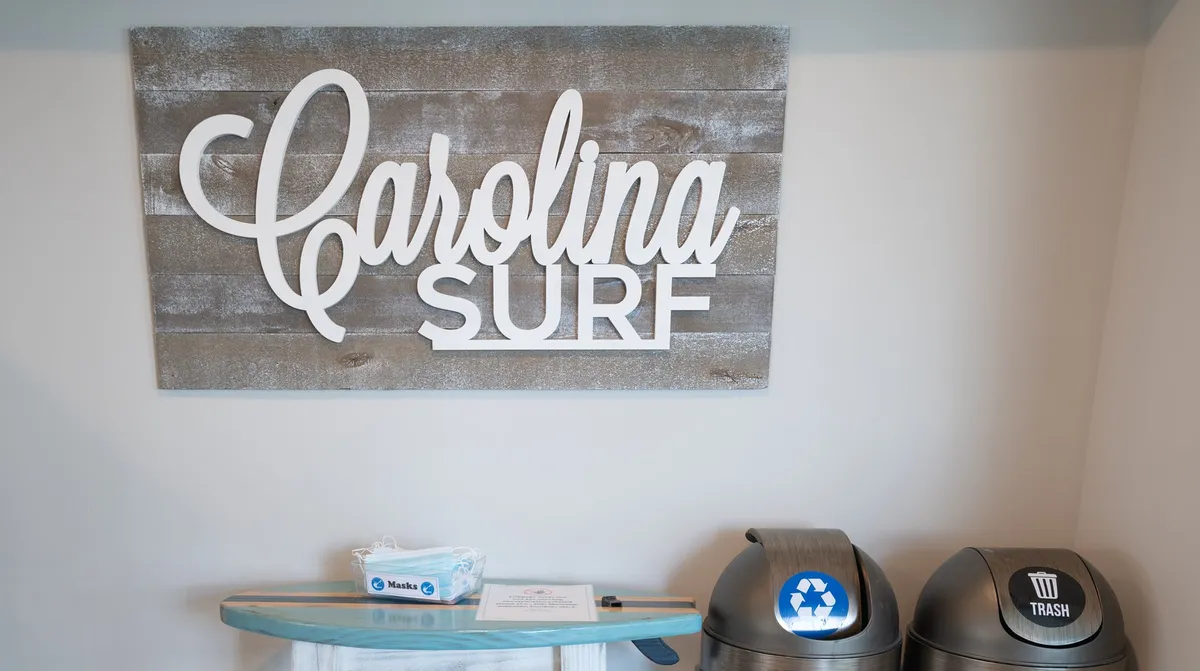 The 'Carolina Surf' sign is visible above a recycling and trash bin area, with cleaning supplies nearby. The setting gives a clear nod to the property's localized character and a welcoming, community-focused atmosphere.