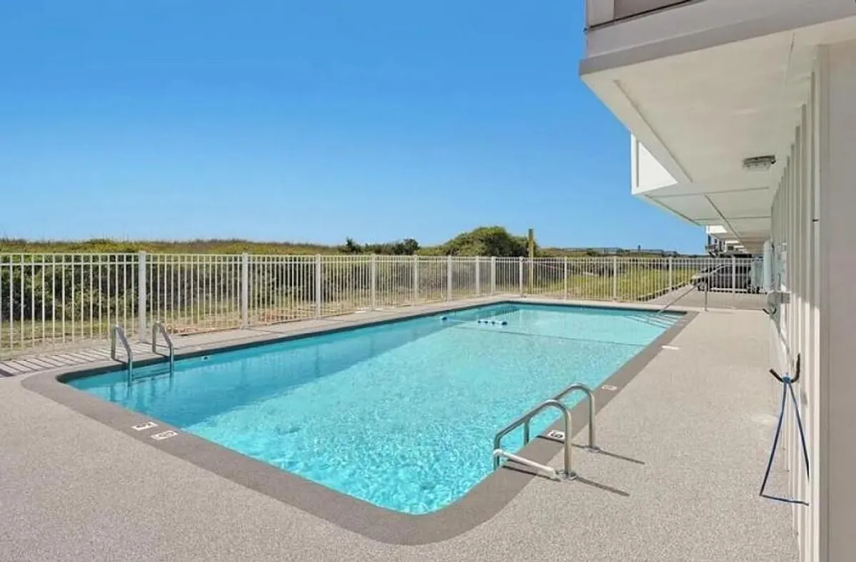 A sparkling outdoor swimming pool surrounded by greenery and enclosed by a safety fence. The area is sunlit and inviting, with ample space for families and guests to relax, sunbathe, or enjoy a refreshing dip.