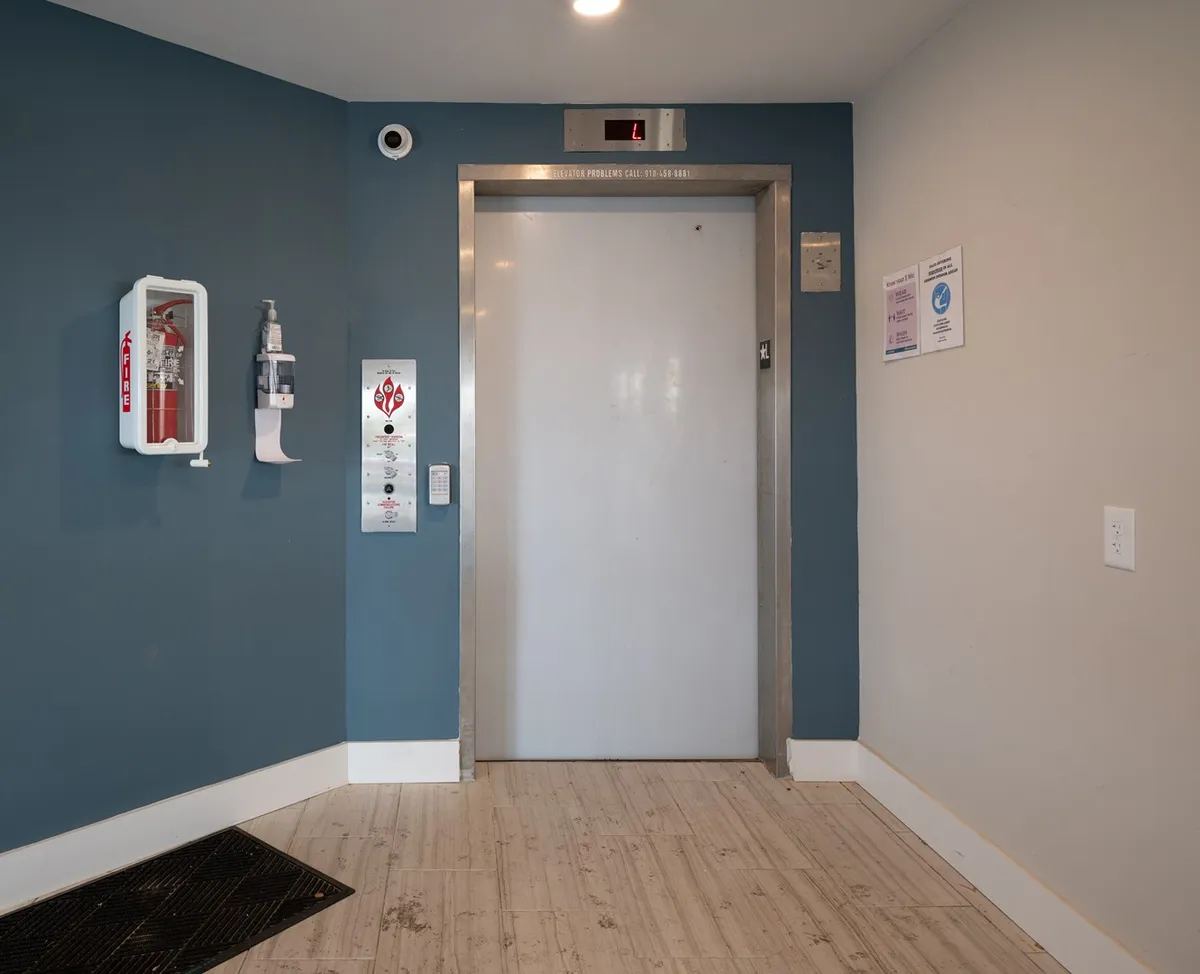 An elevator lobby with a closed elevator door, fire extinguisher, and hand sanitizer station. The space is clean and well-lit, conveying a sense of security and modern convenience.