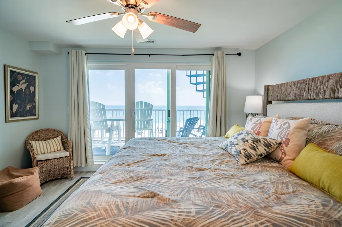 A cozy bedroom features large glass doors opening directly onto the balcony, with colorful pillows on the bed and a wicker chair positioned for enjoying the view. The connection between interior comfort and outdoor scenery is clear, inviting residents to step easily from indoors to out.