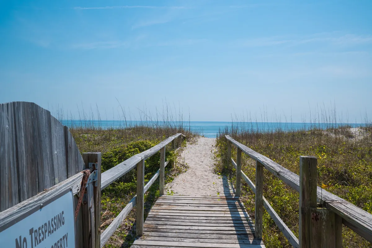 A serene wooden walkway stretches toward the ocean, bordered by coastal vegetation. The view frames the transition from the property to the beach, encapsulating the peaceful yet invigorating experience of oceanfront living.