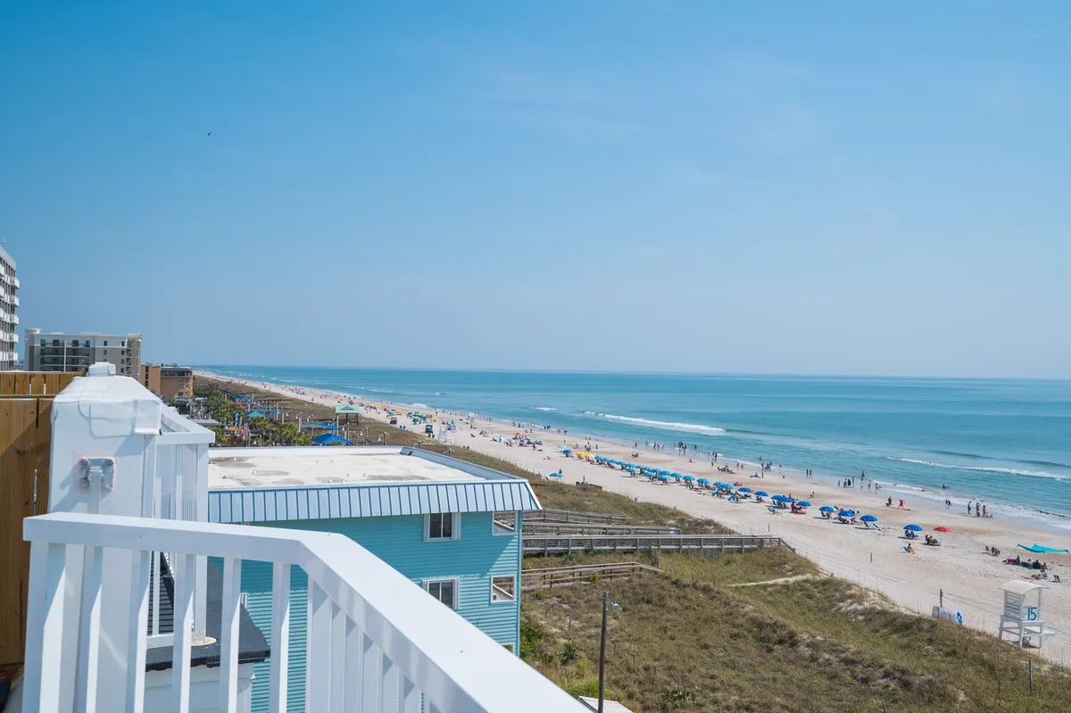 A sun-drenched stretch of beachfront dotted with colorful umbrellas sets a lively scene. Families and groups are seen sunbathing, playing, and strolling along the clear shoreline, with blue skies overhead and gentle waves rolling in. The vibrant, inviting atmosphere instantly communicates classic beach day fun.