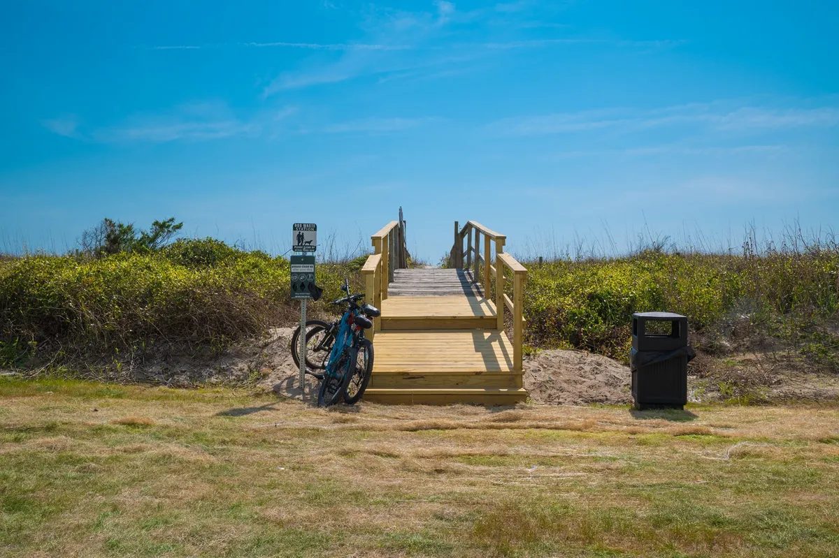 This exterior image features a wooden ramp leading towards the beach, with bicycles parked nearby and beach signage visible. The ocean is just beyond, and the scene suggests easy access for both walkers and bikers, with a relaxed, active vibe.