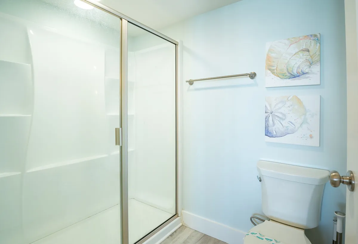 A modern bathroom with a glass shower enclosure, toilet, and shell-themed artwork. The tranquil and clean design highlights the opportunity for relaxation and personal care routines after a day of activities.